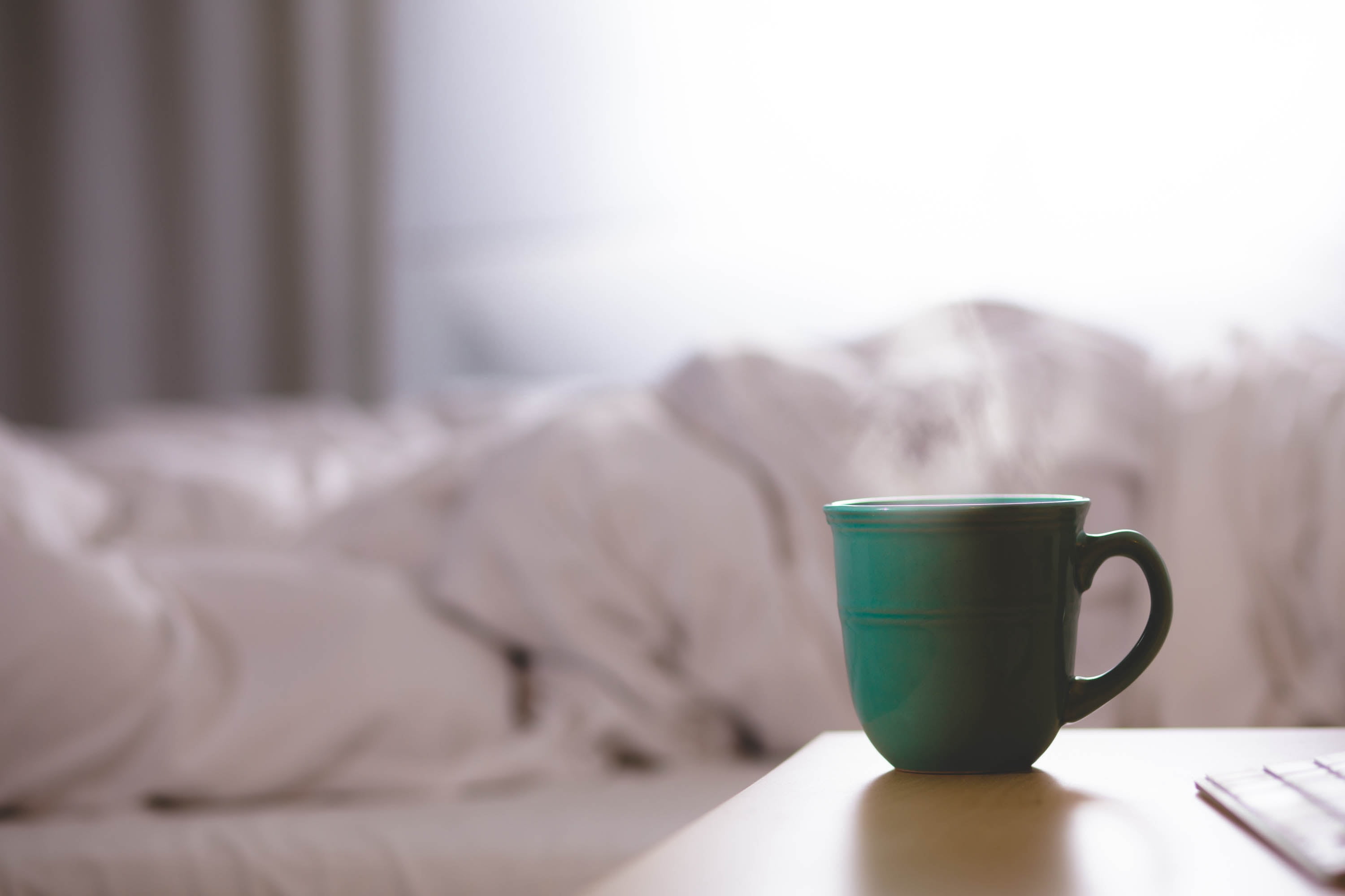 Green steaming teacup on bedside table with big white comforter on bed in background