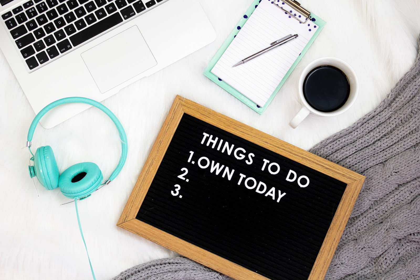 Black sign that says 'Things to do 1. Own Today' beside turquoise headphones and notebook, beside a cup of black coffee and the keyboard of a Macbook